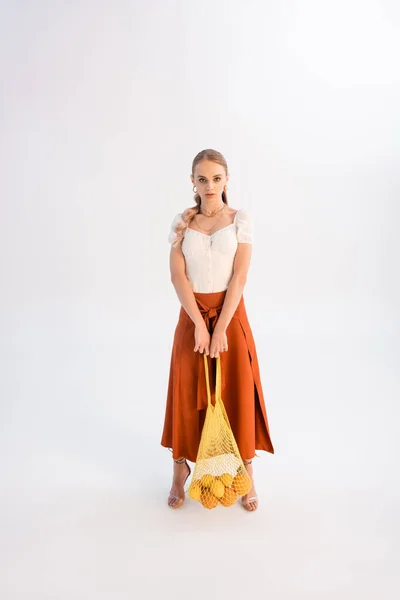 Vue pleine longueur de la femme à la mode posant avec des agrumes dans un sac à ficelle isolé sur blanc — Photo de stock
