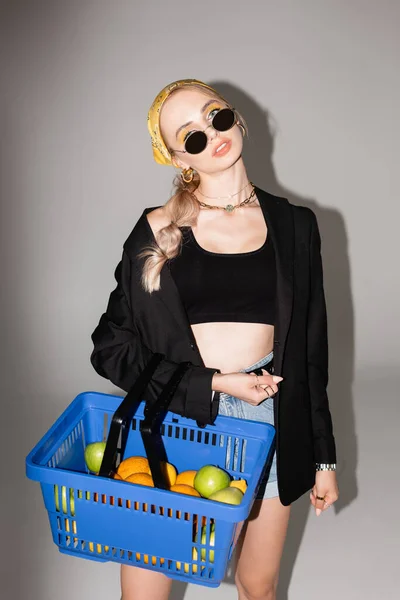 Mujer de moda posando con cesta de la compra azul con frutas sobre fondo gris - foto de stock