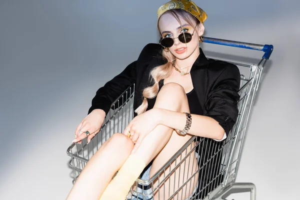 Fashionable woman posing in shopping cart on grey background — Stock Photo