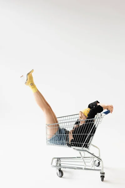 Vista lateral de la mujer de moda en traje negro y amarillo posando en el carrito de la compra en blanco - foto de stock