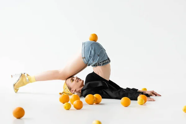 Fashionable woman posing on floor with legs on head near scattered citrus fruits on white background — Stock Photo