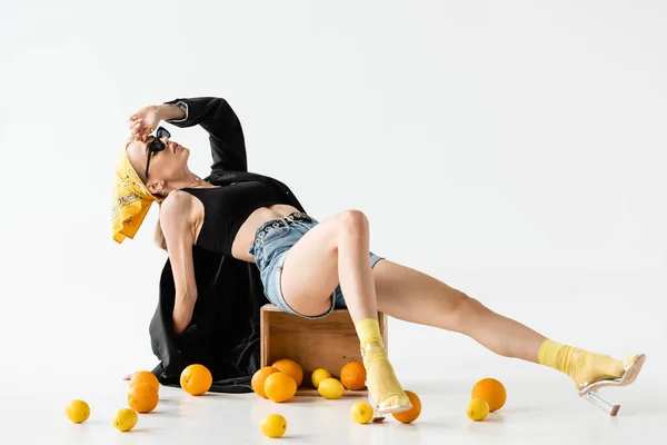 Fashionable woman posing on wooden box near scattered citrus fruits on white background — Stock Photo