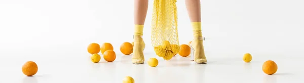 Ausgeschnittene Ansicht weiblicher Beine in gelben Socken und Sandalen und String-Tasche in der Nähe verstreuter Zitrusfrüchte auf weißem Hintergrund, Panoramaaufnahme — Stockfoto