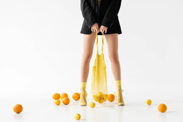 Vista ritagliata della donna alla moda in posa con borsa stringa vicino agli agrumi sparsi su sfondo bianco — Foto stock