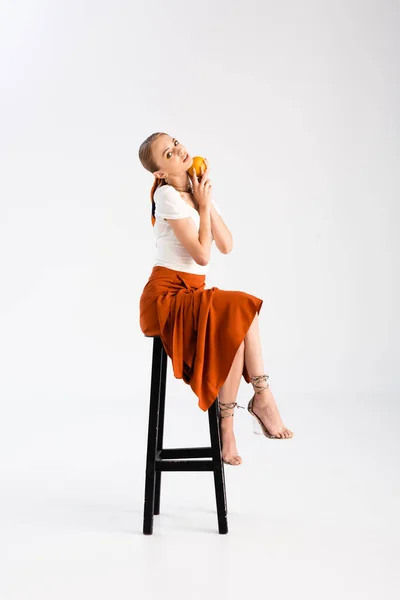 Elegante mujer rubia posando con naranja en silla sobre fondo blanco - foto de stock