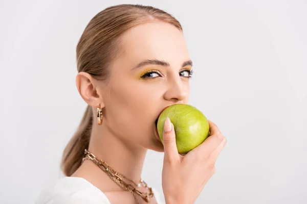 Блондинка кусає зелене яблуко і дивиться на камеру ізольовано на білому — стокове фото