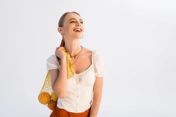 Rustic blonde woman with citrus fruits in yellow string bag laughing isolated on white — Stock Photo