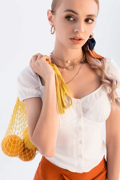 Rustic blonde woman posing with citrus fruits in yellow string bag and looking away isolated on white — Stock Photo