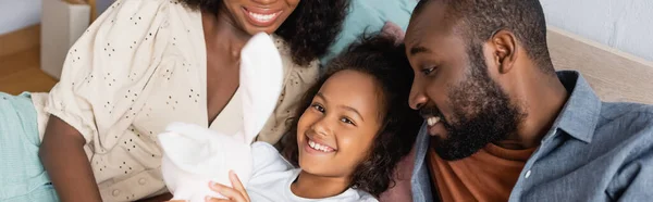 Vista de ángulo alto del niño afroamericano sosteniendo conejito de juguete mientras está acostado en la cama cerca de los padres, imagen horizontal - foto de stock