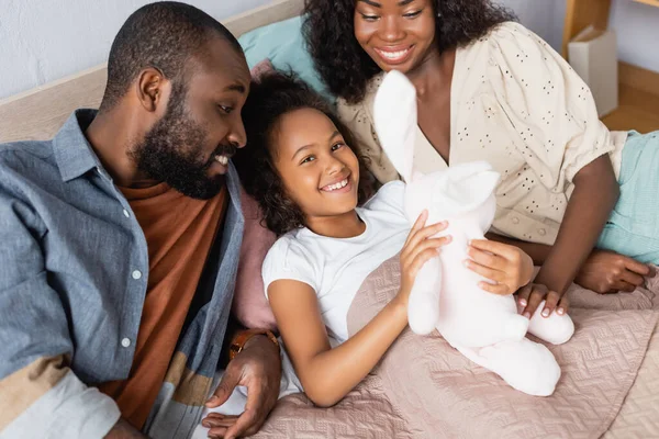 Visão de alto ângulo do jovem casal afro-americano sentado perto da filha deitado sob cobertor e segurando coelho de brinquedo — Fotografia de Stock