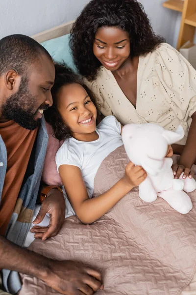 Vista ad alto angolo del bambino afro-americano che guarda la fotocamera mentre giace sotto la coperta e tiene il coniglietto giocattolo vicino ai genitori — Foto stock
