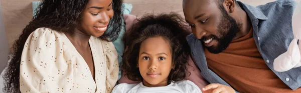 Panoramic crop of african american child lying in bed and looking at camera near parents — Stock Photo