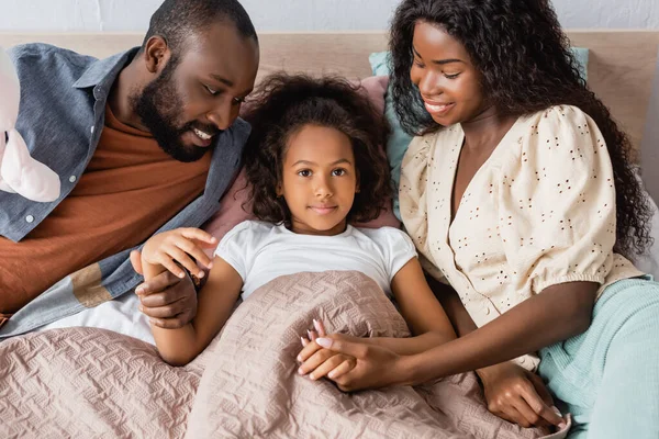 Vista ad alto angolo di africana ragazza americana sdraiata a letto e guardando la fotocamera mentre i genitori seduti vicino e toccare le mani — Foto stock