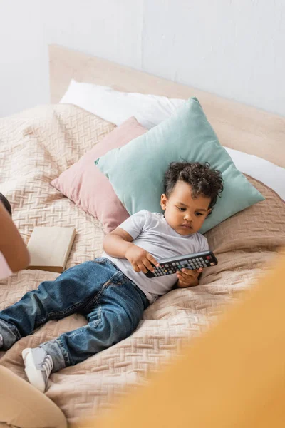 Vista de ángulo alto de niño afroamericano en camiseta blanca y jeans que sostienen el control remoto de la televisión mientras están acostados en la cama - foto de stock