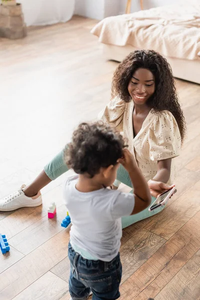 Visão de alto ângulo da mulher afro-americana sentada no chão e mostrando smartphone para filho pequeno — Fotografia de Stock