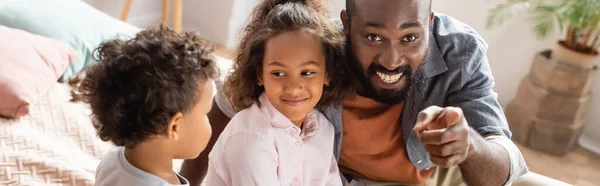 Horizontal image of african american man pointing with finger near kids at home — Stock Photo