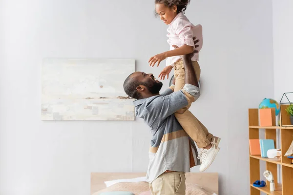 Side view of young african american man raising daughter above head while having fun at home — Stock Photo