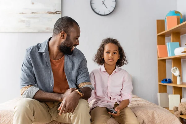Jovem homem americano africano em roupas casuais sentado perto da filha segurando tv controlador remoto — Fotografia de Stock