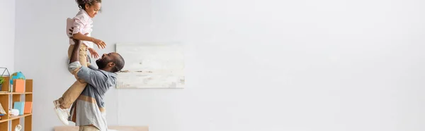 Imagem horizontal do pai afro-americano se divertindo enquanto segurava a filha acima da cabeça em casa — Fotografia de Stock