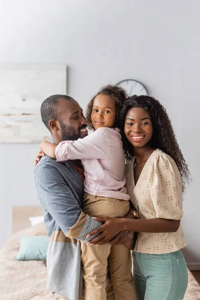 African american man holding daughter on hands while mother hugging them and looking at camera — Stock Photo