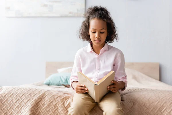 Concentrated african american girl in white shirt reading book while sitting on bed — Stock Photo