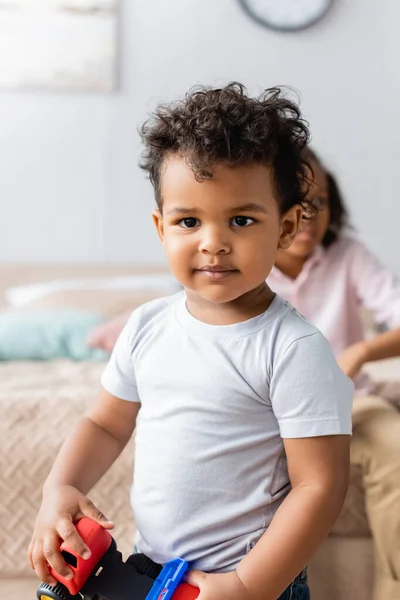 Enfoque selectivo de niño afroamericano en camiseta blanca mirando a la cámara cerca de la hermana sentada en la cama - foto de stock