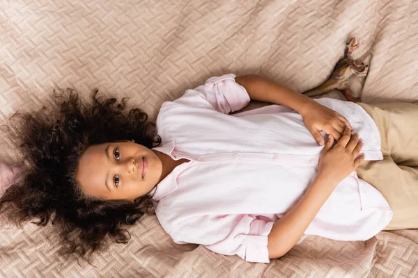 Vista superior da menina americana africana em camisa branca descansando na cama enquanto olha para a câmera — Fotografia de Stock