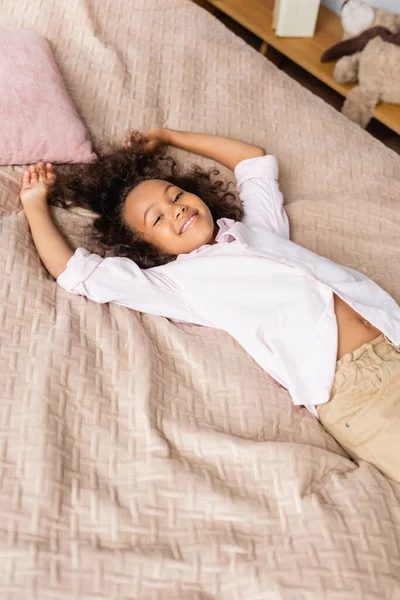 Visão de alto ângulo da menina americana africana em camisa branca deitada na cama e olhando para a câmera — Fotografia de Stock