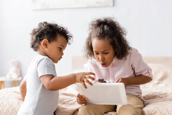 Afrikanische Amerikanerin überrascht mit digitalem Tablet in der Nähe ihres kleinen Bruders im Schlafzimmer — Stockfoto