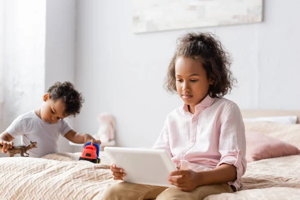 Concentrato africano americano ragazza utilizzando digitale tablet vicino fratello giocare con giocattoli in camera da letto — Foto stock
