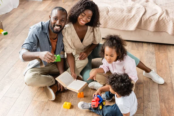 Visão de alto ângulo da família afro-americana sentada no chão com blocos de construção e livro — Fotografia de Stock