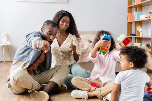 Homem americano africano apontando com o dedo perto da esposa e crianças brincando com blocos de construção no chão — Fotografia de Stock