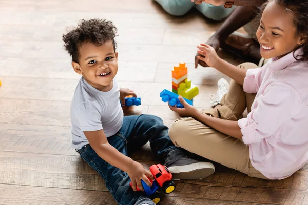 Vista de ángulo alto de hermano y hermana afroamericanos jugando con camión de juguete y bloques de construcción en el piso en casa - foto de stock