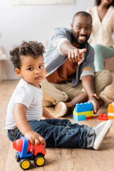 Foco seletivo do menino afro-americano sentado no chão com caminhão de brinquedo perto pai animado apontando com o dedo — Fotografia de Stock