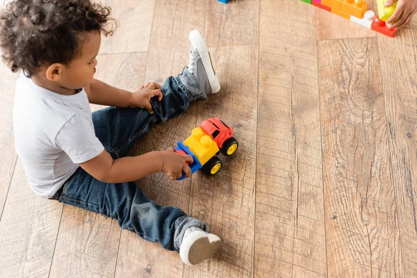 Вид зверху на афроамериканського хлопчика в джинсах, який грає з іграшковою вантажівкою на дерев'яній підлозі — стокове фото