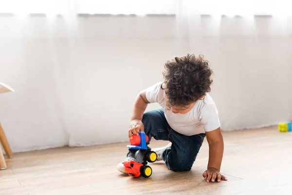 Кудрявый африканский мальчик играет с игрушечным грузовиком на деревянном полу дома — стоковое фото