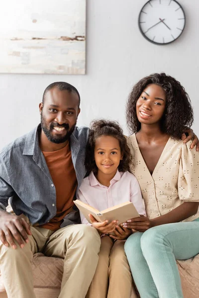 Молоді афроамериканські батьки і дитина з книгою, дивлячись на камеру, сидячи вдома — стокове фото