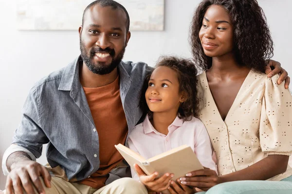 Joven afroamericano hombre tocando hombro de esposa mientras sentado cerca hija sosteniendo libro - foto de stock