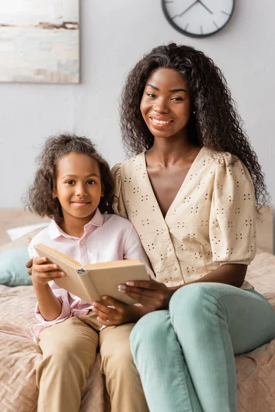 Afro-americana mãe e filha olhando para a câmera enquanto lendo livro juntos — Fotografia de Stock