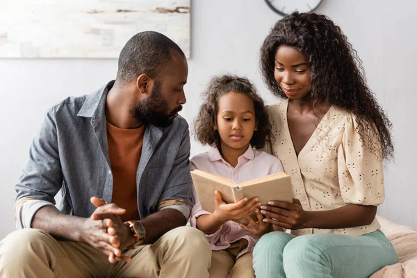 Joven afroamericano marido y esposa sentado cerca concentrado hija lectura libro - foto de stock