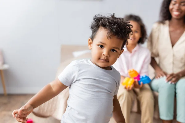 Selektiver Fokus des lockigen afrikanisch-amerikanischen Kindes im weißen T-Shirt, das in die Kamera neben Mutter und Schwester im Schlafzimmer blickt — Stockfoto