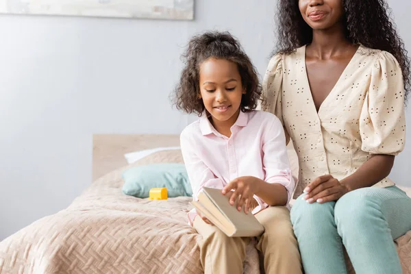 Mujer afroamericana sentada en la cama cerca de la hija sosteniendo libro - foto de stock