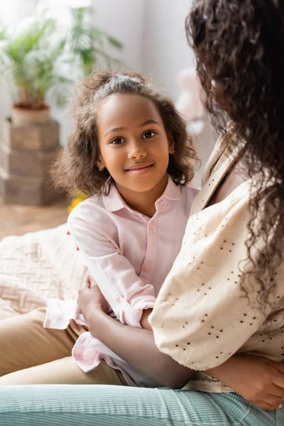 Afrikanisches amerikanisches Mädchen umarmt Mutter, während sie zu Hause in die Kamera schaut — Stockfoto