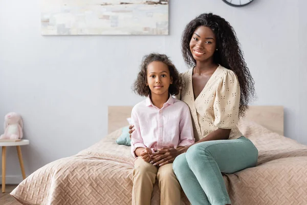 African american mother and daughter in casual clothes looking at camera while sitting on bed — Stock Photo