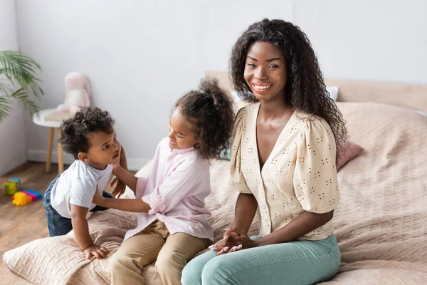 Joven afroamericana mujer mirando a cámara cerca hija e hijo en dormitorio - foto de stock