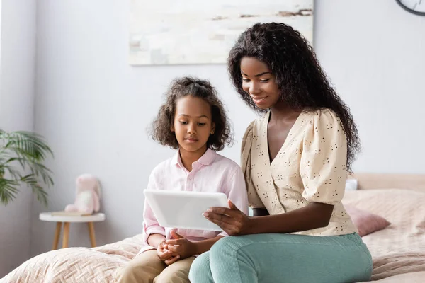 Joven afroamericana mujer usando digital tablet cerca hija en dormitorio - foto de stock