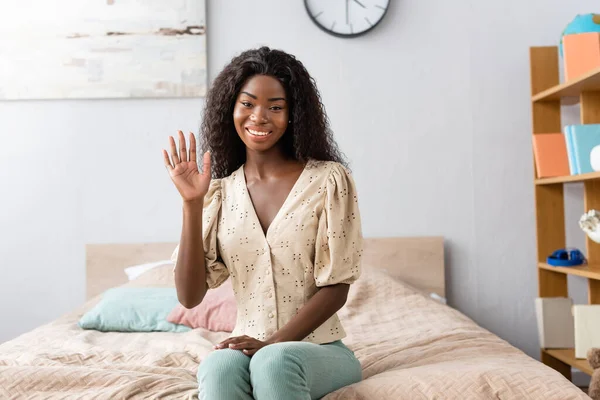Junge afrikanisch-amerikanische Frau in Bluse und Hose sitzt auf dem Bett und winkt mit der Hand in die Kamera — Stockfoto