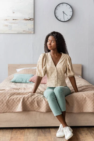 Mujer afroamericana pensativa en blusa y pantalones sentados en la cama y mirando hacia otro lado - foto de stock