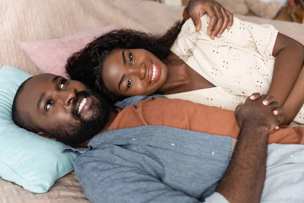 Высокий угол обзора африканского американца, обнимающего жену, смотрящего в камеру, лежащего в постели — стоковое фото