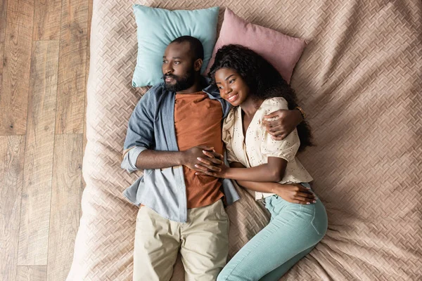 Vue de dessus du mari et de la femme afro-américains tenant la main et regardant loin tout en étant couché dans le lit dans des vêtements décontractés — Photo de stock